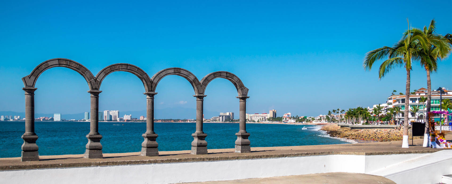 Picture of the Malecon boardwalk in Puerto Vallarta, Mexico.  The pillars of Los Arcos are in the foreground, and is framed by the boardwalk, palm trees and hotels.